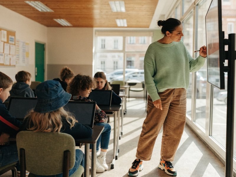Nauczycielka przy ekranie w klasie, uczniowie przy ławkach z tabletami.